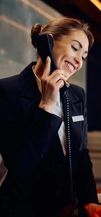 Uśmiechnięta recepcjonistka w eleganckim stroju rozmawiająca przez telefon stacjonarny.